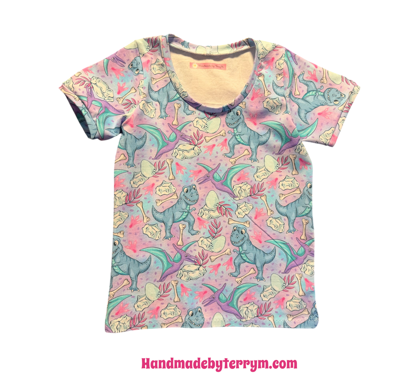 A short-sleeve crewneck t-shirt featuring a colorful cartoon dinosaur print on a light pink background, displayed flat against a white background.